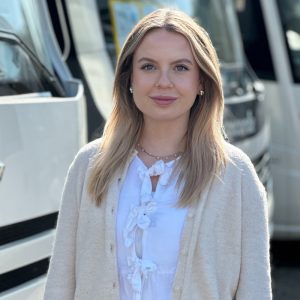 Eine Frau mit blonden Haaren und einem weißen Oberteil steht vor einem Bus.