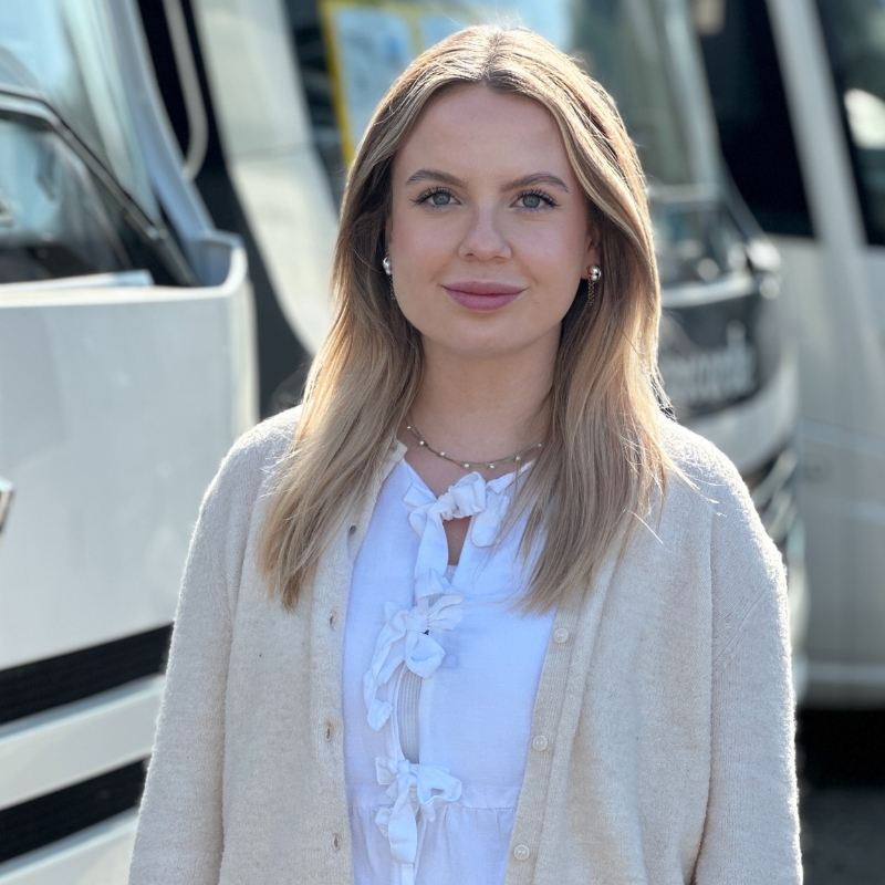 Eine Frau mit blonden Haaren und einem weißen Oberteil steht vor einem Bus.