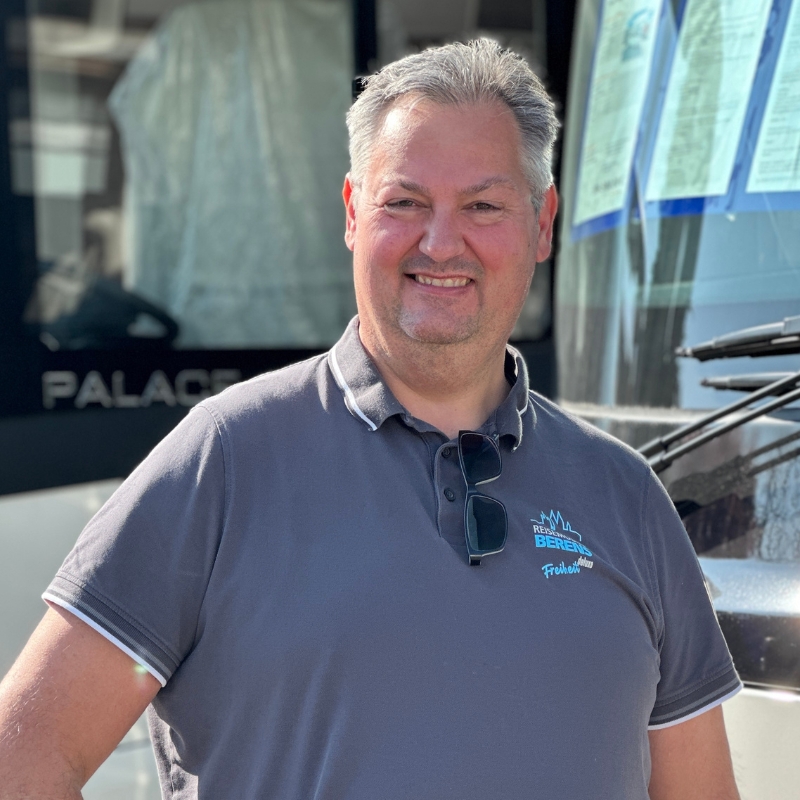 Mann in einem grauen Poloshirt mit Sonnenbrille steht vor einem Fahrzeug.