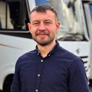 Mann mit kurzem Bart und blauer Hemd steht vor einem weißen Fahrzeug.