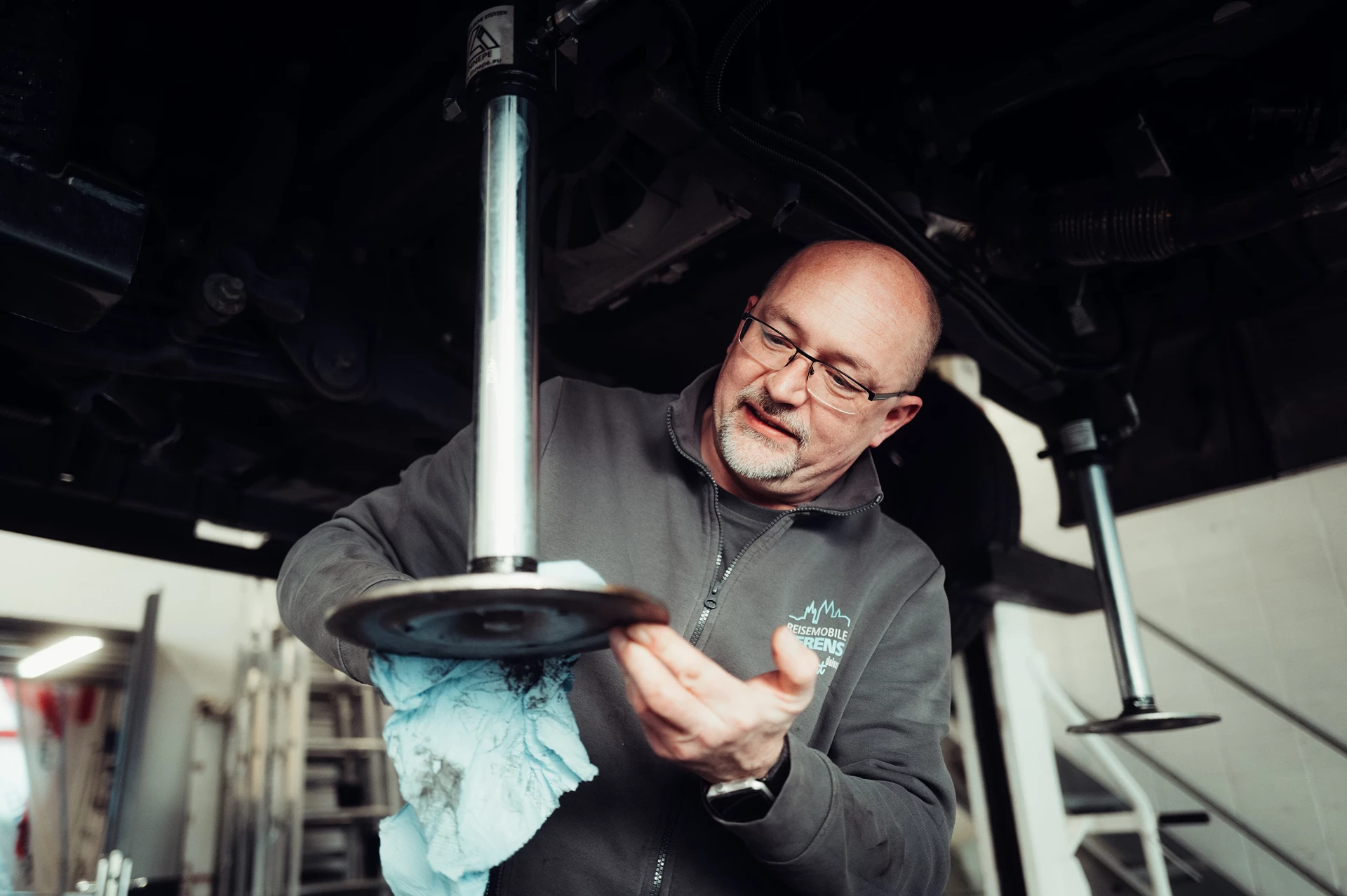 Ein Mechaniker untersucht ein Fahrzeugteil unter einem Auto und hält ein Tuch in der Hand.