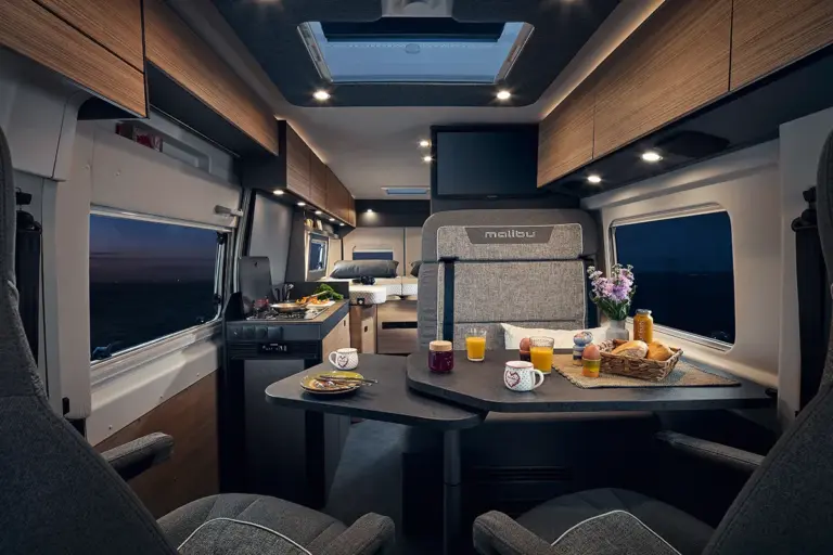 Innenansicht eines Camper Vans mit einem Essbereich, Tischen und Stühlen sowie Küchenzeile.