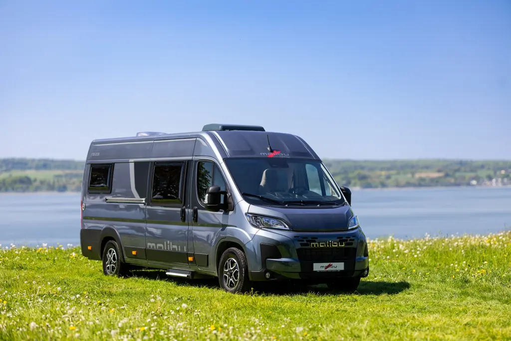 Grauer Malibu Relax Camper Van steht auf einer Wiese mit Wasser im Hintergrund.