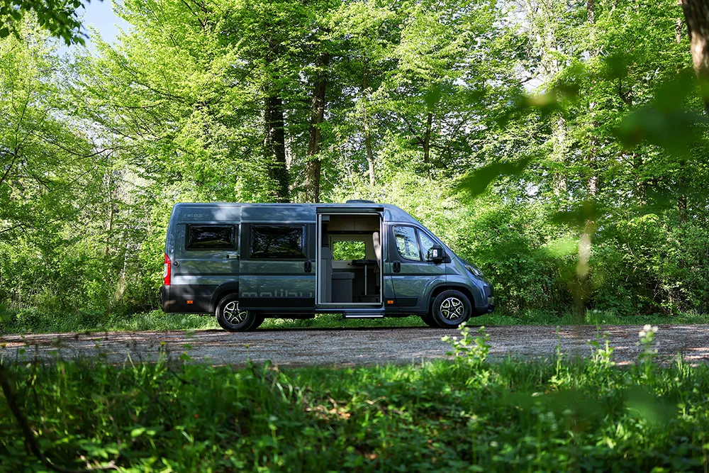 Graues Malibu Wohnmobil mit geöffneter Tür, abgestellt im Wald.