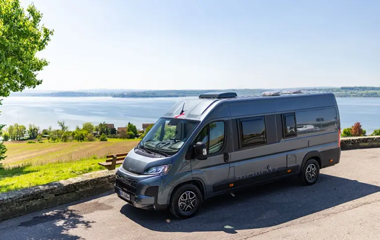 Grauer Camper Van parkt an einem Aussichtspunkt mit Blick auf einen See und die umliegende Landschaft.