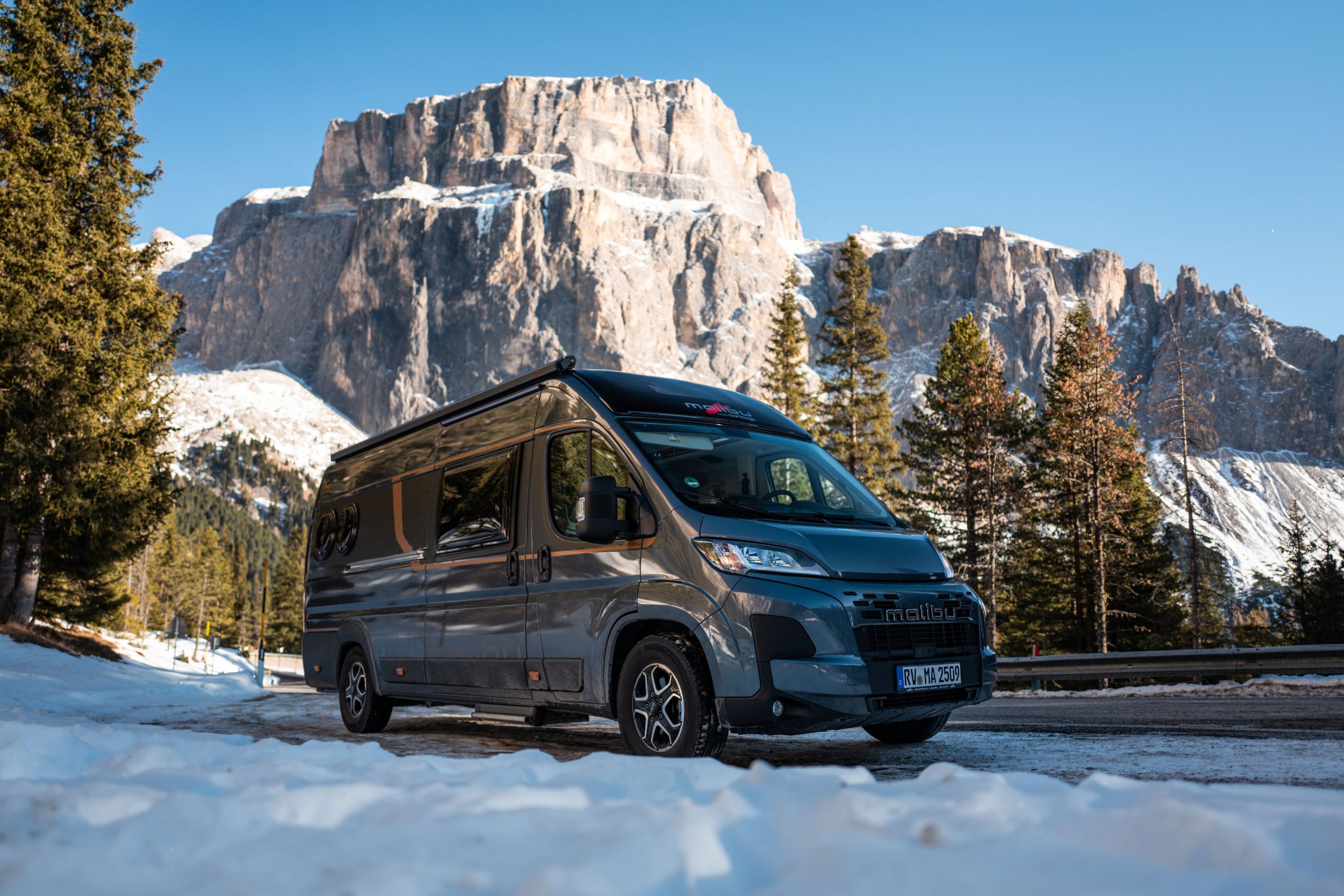 Graues Wohnmobil von Malibu steht auf einer schneebedeckten Straße mit Bergen im Hintergrund.